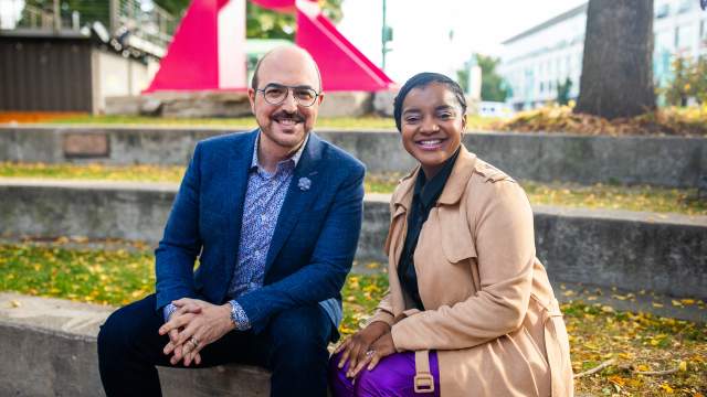 Ashley Jordan and David Caruso sit together smiling in an outdoor space with a large pink sculpture in the background