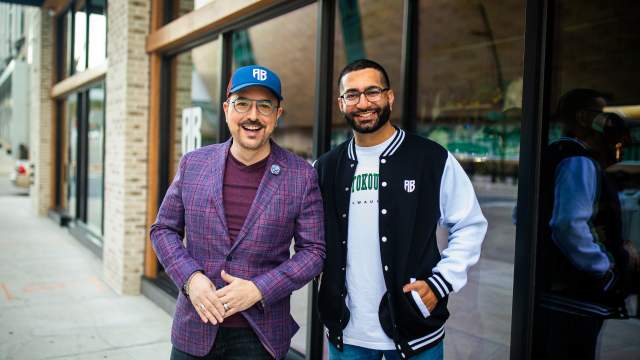 Two men stand smiling together outside a modern commercial building with large glass windows. On the left, a man wears a purple plaid blazer over a burgundy shirt with a blue "AB" cap and glasses. On the right, Nimesh "Nemo" Ruparel wears his signature black and white varsity jacket with "MILWAUKEE" text in green letters and "AB" logo, paired with glasses and a beard. The setting appears to be an urban commercial district with contemporary architecture featuring brick and glass storefronts.