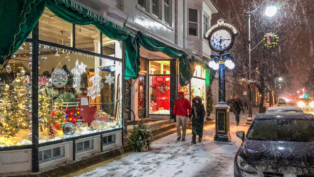 People walking along a snow-covered downtown street at night, passing festive shop windows decorated with holiday lights and ornaments.
