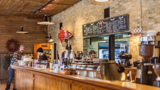 Interior of a cozy coffee shop with a long wooden counter, baristas preparing drinks, and a large chalkboard menu on a brick wall.