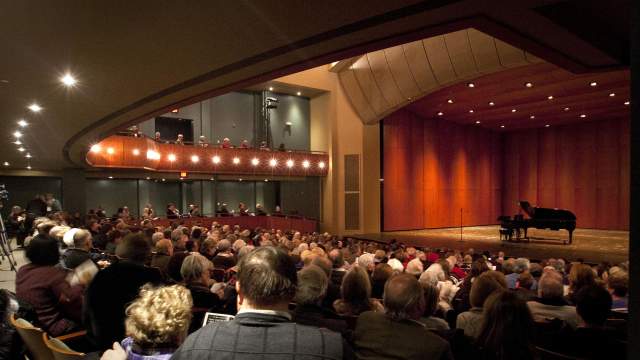 Audience seated in a concert hall watching a piano performance on stage under warm lighting.