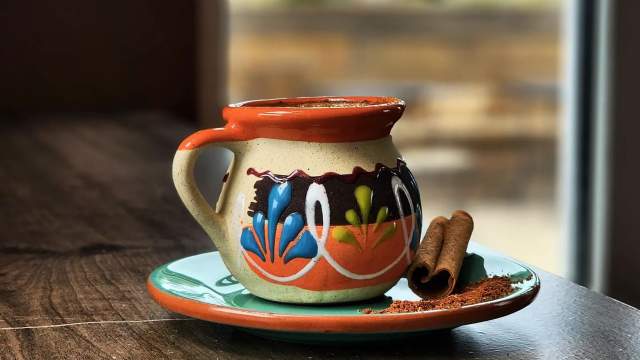 A colorful ceramic mug with a painted floral design on a matching saucer, served with two cinnamon sticks on a wooden table near a window.