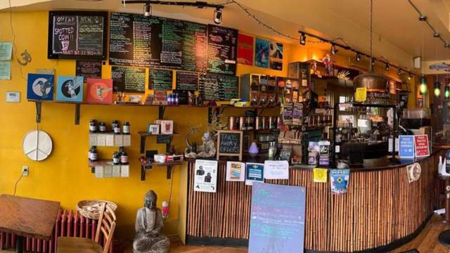 Colorful, cozy café interior with a bamboo-covered counter, handwritten chalkboard menus on a bright yellow wall, eclectic artwork, and a small Buddha statue near the seating area.