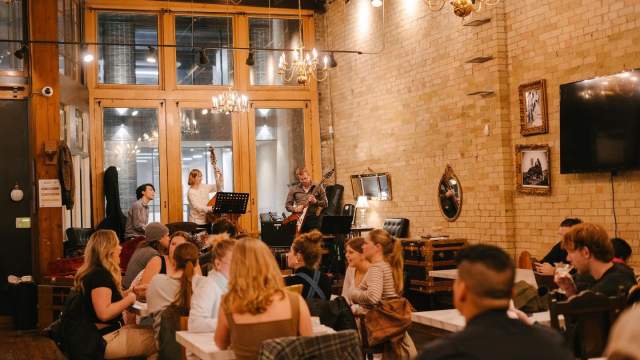 Warm, inviting restaurant or café interior with exposed brick walls, chandeliers, and large windows, where groups of people are seated at tables dining and chatting.