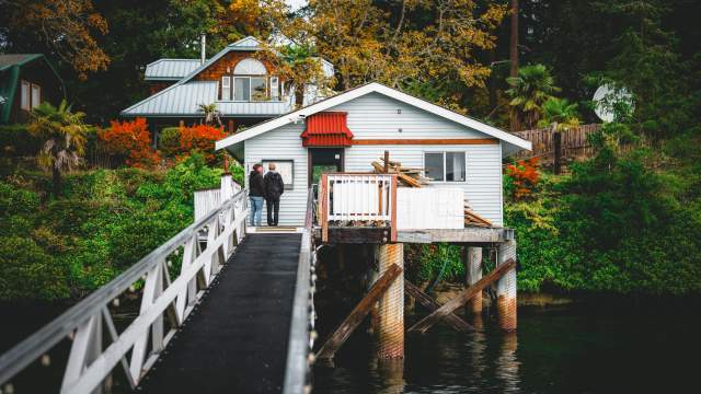Two people stand on a narrow walkway leading to a small oceanfront house on stilts above the water, surrounded by trees and fall foliage.