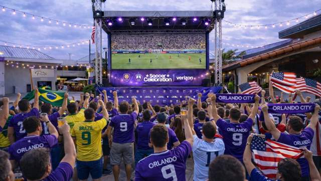 Renderings of viewing areas, Outdoor watch party with a crowd gathered in front of a large elevated screen displaying a soccer match. Attendees wear team jerseys and hold flags while standing beneath string lights and stage lighting in an open-air venue.