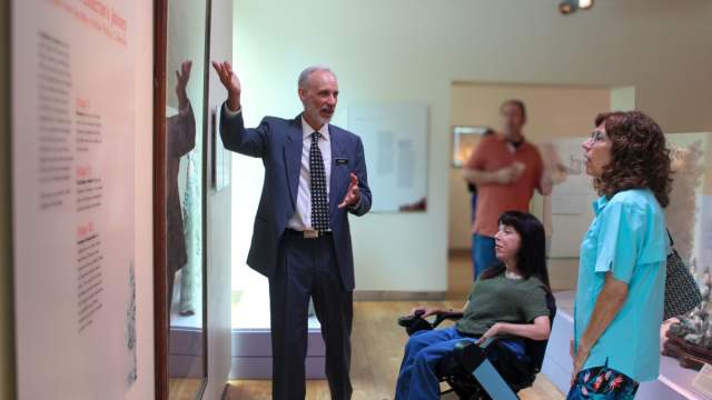 Man pointing at a display in a museum with a woman in a wheel chair, a woman standing,  and a man all watching and listening intently.