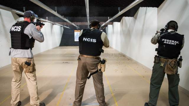 3 men pointing away from the camera, all with black vests on. The 2 men on the left have vests that say "NCIS" and the man on the right has "federal agent" written on the back of his vest. They are holding guns at an indoor gun range.