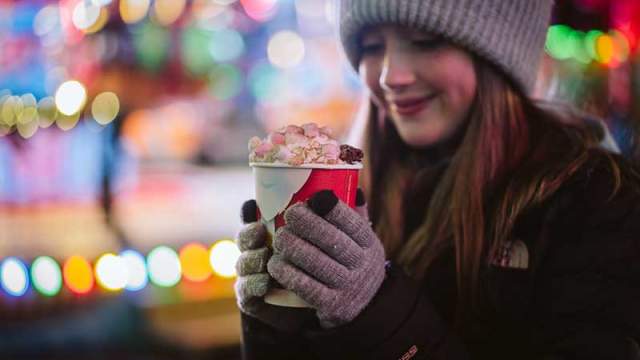 A girl in a hat and gloves holding a hot chocolate covered in marshmallows.