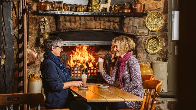 Two women in front of a pub fire drinking hot chocolate at a table.