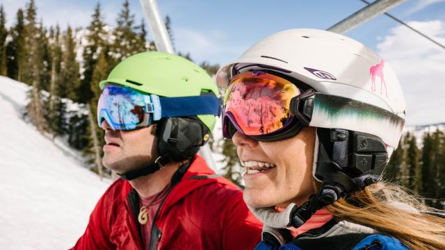 A man and a woman wearing helmets and goggles riding the ski lift at Salt Lake's ski resorts