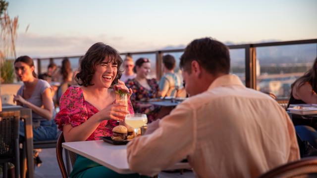 A man and a woman having dinner at a table. The woman is smiling and laughing. The background is a sunset shot over Salt Lake