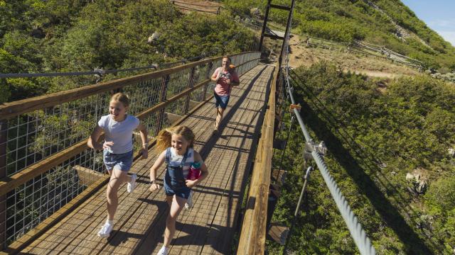 Father and two girls running across a walking path suspension bridge with green mountainside in the background