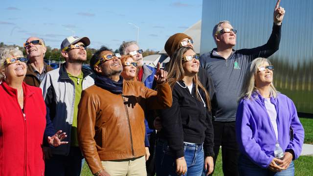 A group of individuals, wearing solar eclipse glasses and looking to the sky, as if they are viewing a solar elcipse.