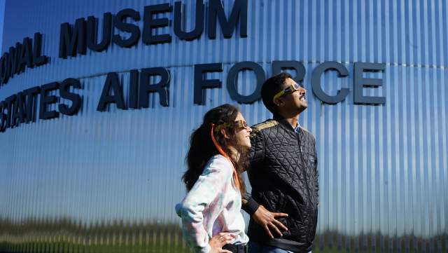 A man and a teenage girl, both wearing eclipse glasses and standing in front of the National Museum of the U.S. Air Force, looking skyward to the right, as if viewing a solar eclipse.