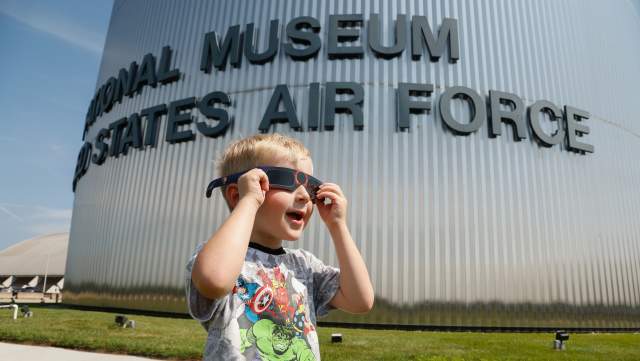 A young boy wearing solar eclipse viewing glasses, standing in front of the National Museum of the U.S. Air Force