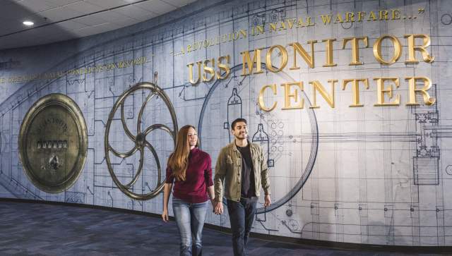 A couple walks in the Mariners Museum, behind them is a sign that reads "USS Monitor Center"