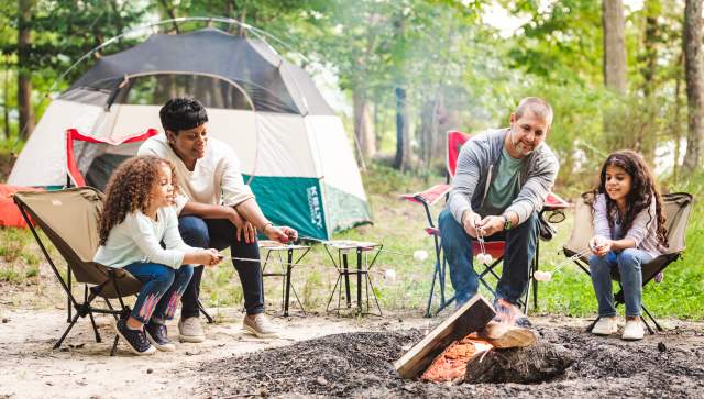 Family of 4 roasts marshmallows over a fire in front of a tent in the woods