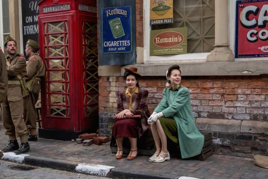 Actors dressed in 1940s clothing sit beside a vintage British red telephone box on a cobbled street, surrounded by period military uniforms and retro advertising signs.