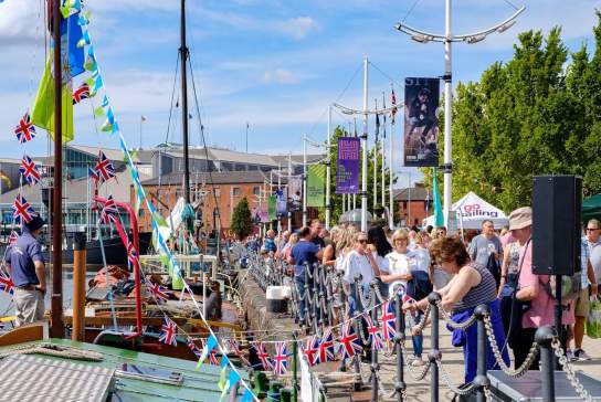 Crowds of visitors walking along the waterfront promenade at Hull Marina during the Folk and Maritime Music Festival. Traditional boats are moored along the marina in the foreground, decorated with strings of Union Jack bunting and colourful maritime flags. People gather beside the railings to watch the activity on the water and along the quay, while festival banners hang from modern lampposts lining the walkway. Market stalls and event tents can be seen further along the marina, creating a lively festival atmosphere against a backdrop of historic brick warehouses and modern buildings under a bright blue summer sky.