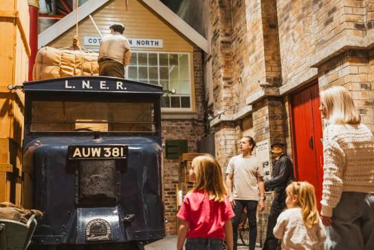 Families exploring a historic railway-themed gallery, looking at an L.N.E.R. vintage delivery truck, luggage display, and recreated station setting inside a Streetlife Museum Hull .