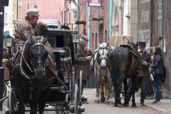 Hullywood film set featuring a vintage horse‑drawn carriage on a narrow cobbled street, with actors in period costumes preparing horses beside historic brick buildings, creating an authentic old‑world production atmosphere.