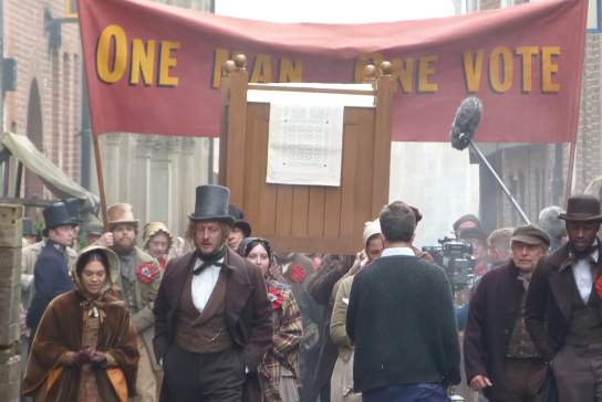 Hullywood film set recreating a historic street protest scene, featuring actors in period costumes marching beneath a large banner reading “One Man One Vote,” surrounded by vintage props, market stalls, and production equipment to create an authentic nineteenth‑century atmosphere.