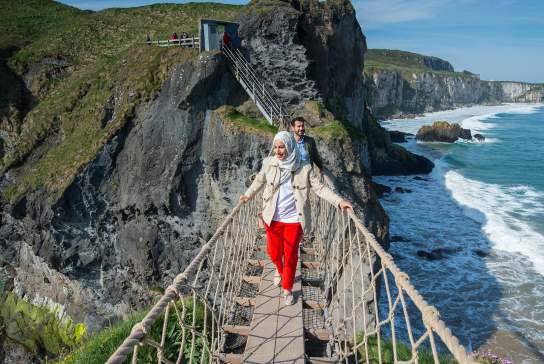 Stunning views of Carrick-a-Rede Rope Bridge, along the Causeway Coastal Route as a couple cross the bridge.