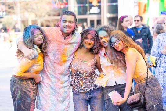 5 people with their arms around each other covered in colourful powder smiling at the Holi event in Guildhall Square