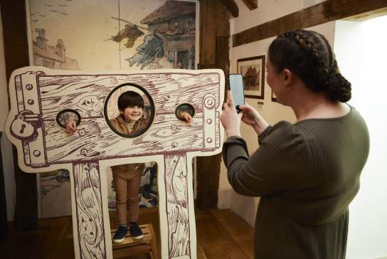 Young boy posing in cardboard cut out of medieval stocks while mother takes photo at Tudor House and Garden