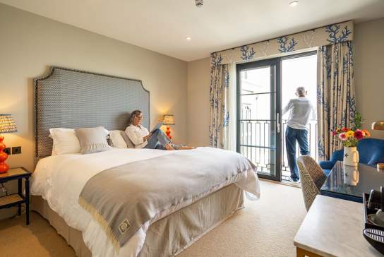 A man and a woman in a hotel room. Woman reading on the bed, man looking out of the window.