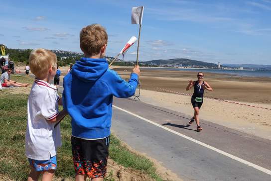 Two young boys cheering on their mum running along the race course waving welsh flags