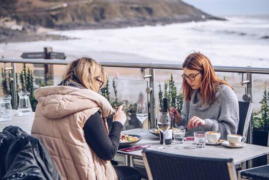 Two women eating outside a restaurant with coast in background