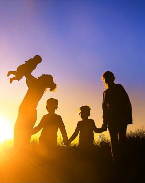 A family is silhouetted against the sun. A mother holds a baby in the air, while three other children hold hands next to them