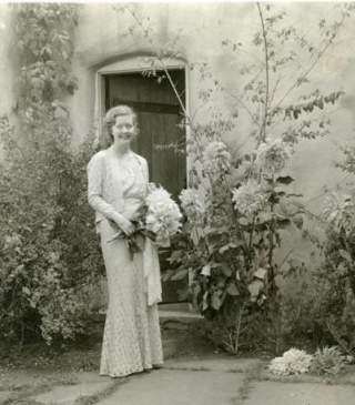 Old film photo of a woman holding a bouquet in front of an adobe building with flowers and vines growing in front of it.