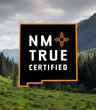 A logo in the shape of the state of New Mexico with text reading NM True Certified in front of a forested area with cloudy skies