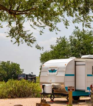 A retro-style camper trailer with blue and white stripes is parked at a campground. Nearby, another RV is visible, surrounded by lush greenery. A tree branch frames the top edge of the scene.