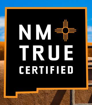 A logo in the shape of the state of New Mexico that read NM True Certified in front of an adobe building with a wooden ladder