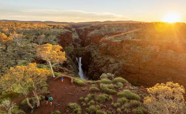 A family walks towards the Joffre Gorge Lookout in Karijini National Park - a waterfall is visible from the lookout and it's surrounded by red rocks, spinifex and snappy gum trees.