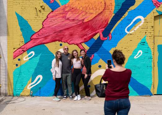 A group of four people poses in front of a vibrant mural featuring a pink bird and abstract shapes, while one person takes a photo.