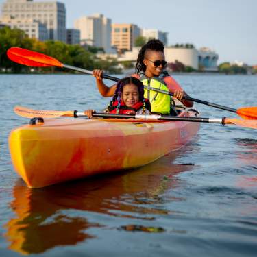 A Black woman and young Black girl kayak on Lake Mendota with the Madison skyline in the background