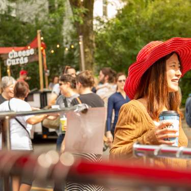 An Asian woman in a large red hat walks through the crowded Willy Street Fair holding a coffee cup.