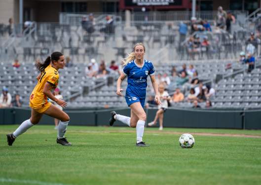 Two female soccer players, one in yellow and the other in blue, compete for the ball on a lush green field, with spectators in the background.