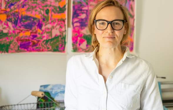 Woman smiles and stands in front of completed artwork hanging on the wall. She is wearing a white shirt and black-framed glasses.