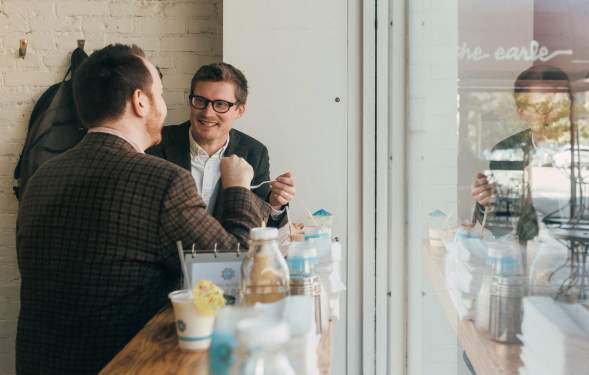 Two men, both wearing dark suits, dine at a windowside counter. They are engaged in conversation.