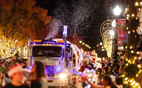 A photo of the Christmas Under The Stars Parade in 2023 featuring a huge float being pulled by an 18-wheeler down Center Avenue. Crowds are seen up and down the street on both sides under the christmas lights and decorations of Center Avenue.