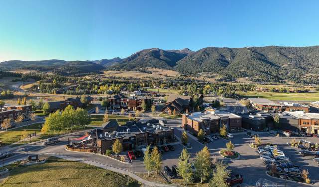 Aerial view of a vibrant mountain town with shops and restaurants, surrounded by lush greenery and rolling hills under a clear blue sky.