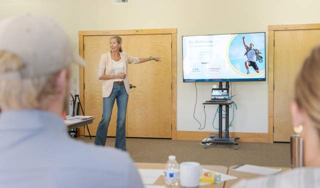 A woman presents in a bright room, gesturing towards a screen displaying a diagram. Attendees are seated at tables, engaged.