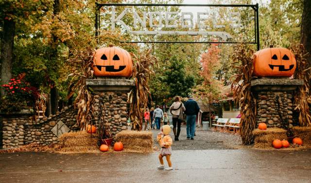 A toddler in front of a large metal sign that reads Knoebels. Two jack o lanterns sit on either side of the sign.