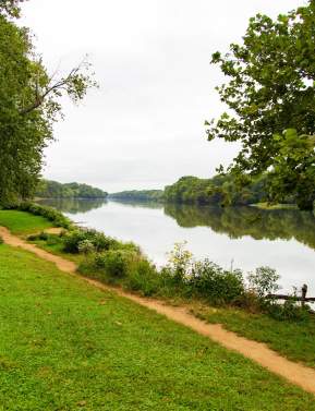 A dirt trail along the Potomac River shoreline at Algonkian Regional Park in Loudoun County, Virginia, with a person fishing in the distance
